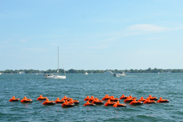 two small boats on Lake Ontario, Toronto harbour, pass by the art installation S O S or Safety Orange Swimmers