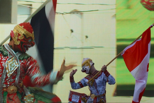 a man in Thai costume and holding a Canadian flag does a Thai traditional dance on the stage at Yonge Dundas Square. behind him a video of another dancer is playing on a large screen 