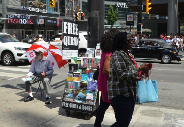 two black women walk past a man sitting in a chair with a large red and white Canadian flag umbrella. he is giving away free quran books on the sidewalk by Yonge street, traffic passing by on the street behind them 