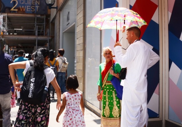a man in long white robes holds an umbrella as he stands on the sidewalk talking to a woman in Southeast Asian clothing 