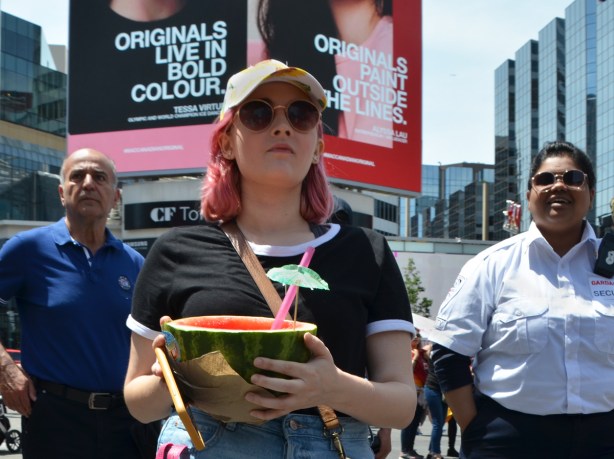 a woman weth pinkish hair carried a half watermelon with a straw and a little green paper umbrella in it, she is the middle of three people standing in Yonge Dundas square 