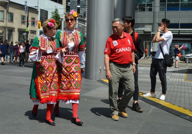two womenin national costume, or traditional clothes of another country, walk through Dundas Square, an Asian man is looking at them with a strange look on his face 