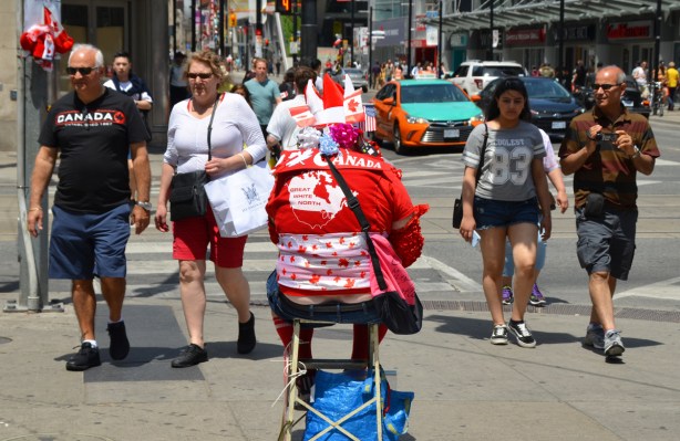 four people walk past a man sitting on a stool, all dressed in red and white with flags and Canadiana