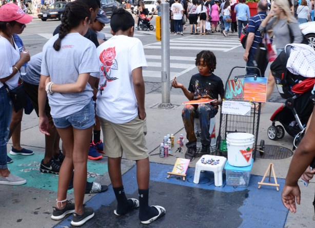 a young black boy sits on a chair at Yonge and Dundas and paints small pictures while people stop to watch him