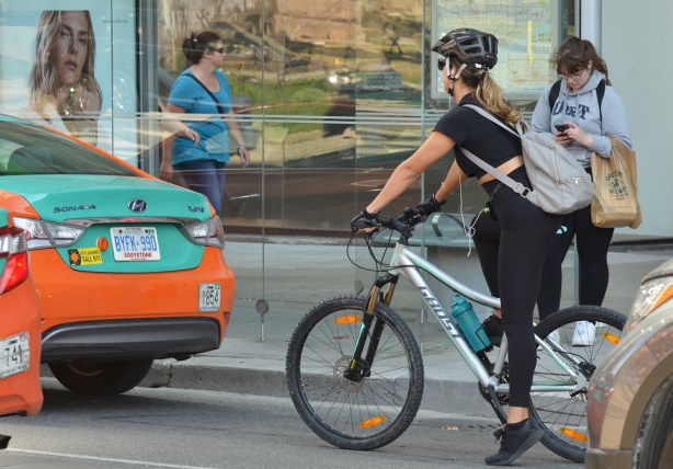 a woman in black on a bike is stopped for a red light in front of a bus shelter. A woman in blue is walking past on the sidewalk, reflections in a store window, a woman in a grey U of T hoodie stands beside the bus shelter. two orange and green taxis are in front of the bike 