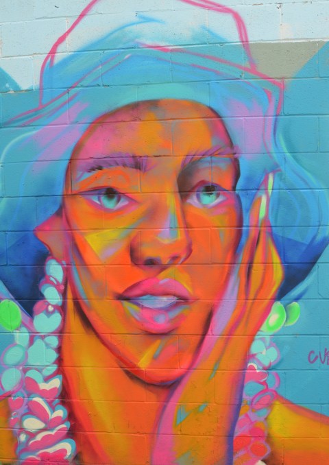 large face mural on an exterior wall in an alley, in bright fluorescent oranges and blues, wearing a large floppy hat 