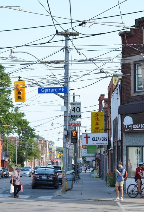 Broadview looking south from Gerrard with utility poles and lots of wires, people crossing the street, some traffic, the clears with the sign with a red cross on it