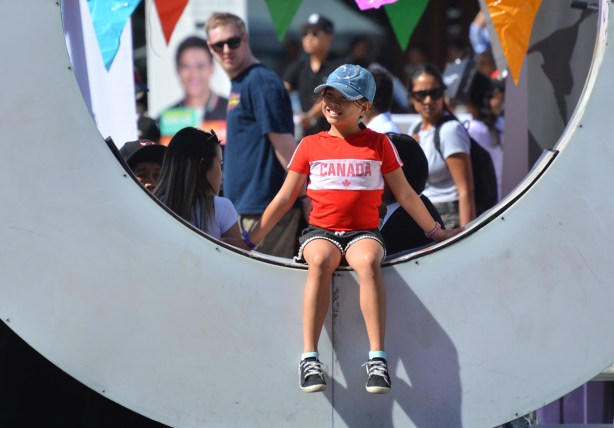 a young girl in a Canada T shirt sits in the O of the 3 D Toronto sign at Nathan Phillips square, people walking behind her, a man turns to look over his shoulder and appears to be looking at the girl 