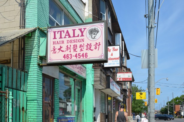 store fronts on Gerrard including one that is painted bright green, signs over the doors including the Italy hair design store with sign in English and Chinese