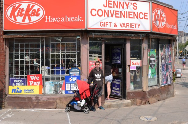 a young man stands beside a stroller outside Jenny's Convenience store on Parliament street, large red and white sign with kit kat logo on it twice - once at each end