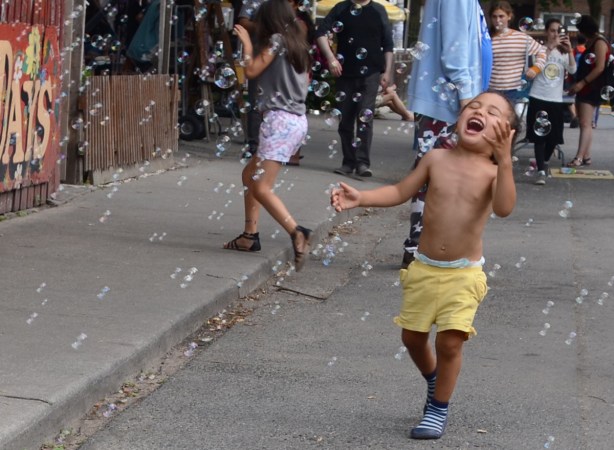 a young boy in yellow shhorts and blue shoes is laughing as he runs through a stream of bubbles, outside on the street 