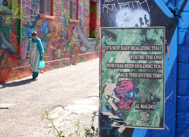 on a wall in Graffiti Alley, a poster with a saying or perhaps poes credited to A.J. Maldo, with a woman in a turquoise sari walking past 