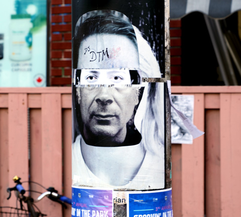 on a utility pole, a paper graffiti piece of a man's face with the eyes from a different picture 