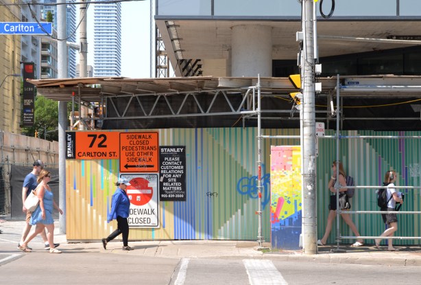 people walking past painted hoardings in front of a construction site, painted in stripes