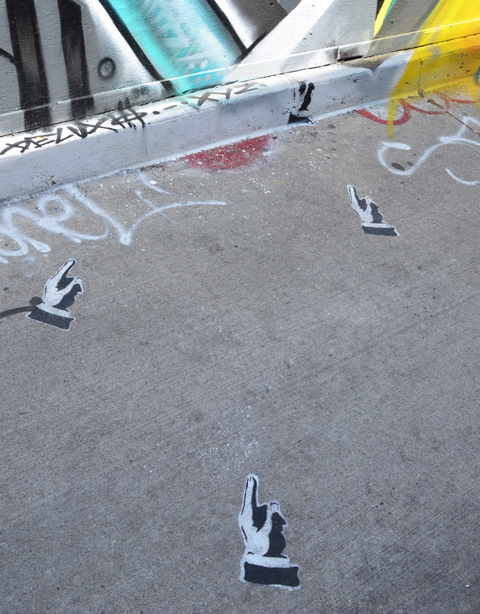 4 black and white stencils of hand and fingers pointing, on the sidewalk, pointing towards a mural on a wall 