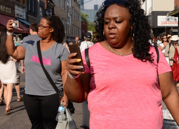 two black women walking down the street while looking at their phones, on in a bright pink T shirt 