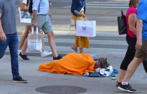 a man sleeps under a bright orange blanket on the sidewalk on Queen Street while people's feet pass by 