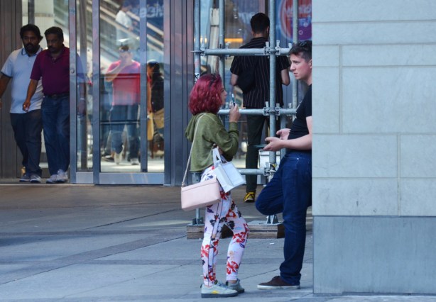 by one of the entrances to the Eaton Centre, outside, a man leans against a wall while a woman with reddish purple hair talks to him. She is wearing floral pants and has a pale pink handbag. Other people are coming out the doors of the Eaton Centre 