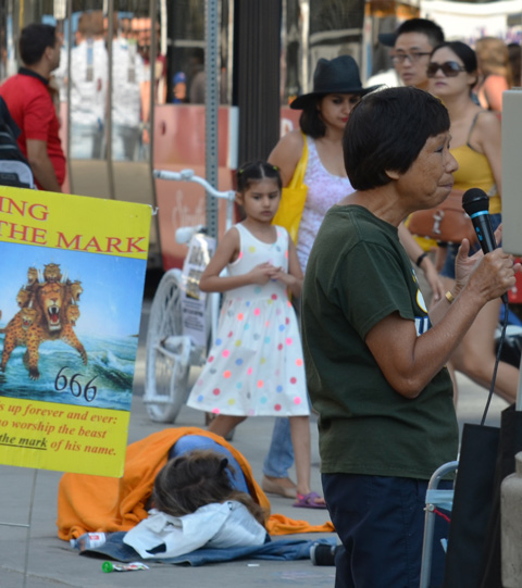 a woman talks into a microphone, about religion, a man sleeps on the sidewalk in the background as people and a TTC streetcar pass by. A mother and daughter are walking on the sidewalk and longing down at the sleeping man 