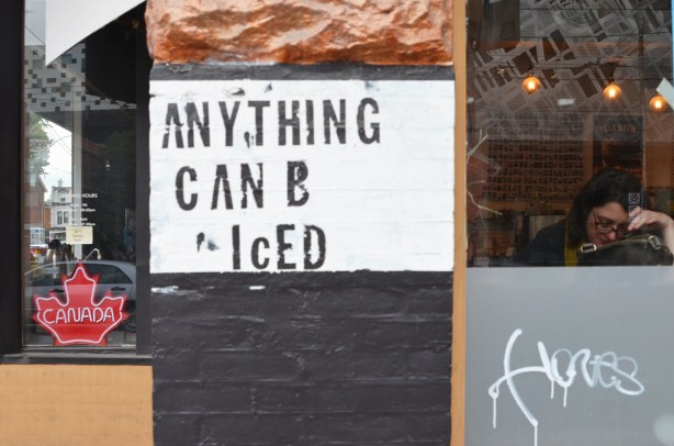 a sign stenciled beside a doorway that says Anything can b iced. Window beside it, with a woman sitting inside by the window 