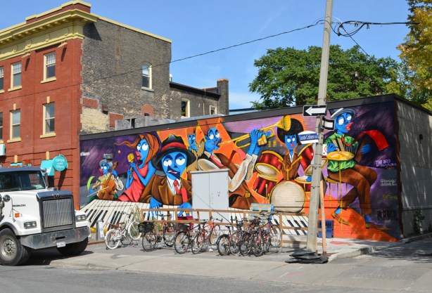 whole mural part of a mural, the band of story tellers, a group of musicians all with blue skin, with bikes in front of it, by Leah Cohen Lane 