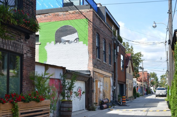 Croft street alley with garages on the left, and apartments above some of them, a mural of a man's face where the bottom half has been painted over with white paint 