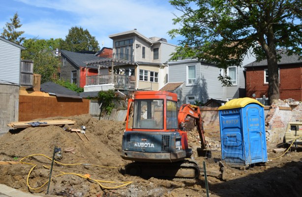 a digger and a blue porta-potty in a vacant lot construction site, with a row of backyards and backs of houses behind