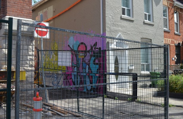 construction fence around a building being built, also a small beige house with white trim. Between the two is an entrance to an alley, there is a mural on the wall of the house, in the alley
