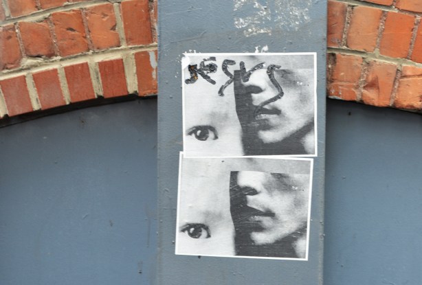 2 paper paste ups on a wall, both are the same, in grey tones, the lower part of a face with one eye beside it 