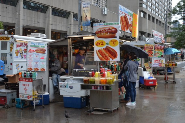 hot dog and sausage vendor on Queen street, woman under umbrella buying something, woman working inside the booth, a man sitting behind, many signs advertising their food 
