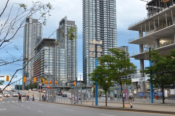 looking south on Spadina near Front, streetcar stop and shelters in the middle of Spadina, some people waiting for streetcars, 2 small trees, tall condos in the background