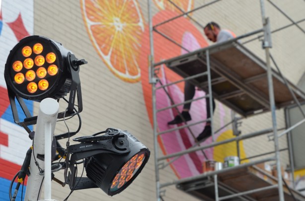 a man sits on scaffolding as he paints a mural, LED spotlights are in the foreground