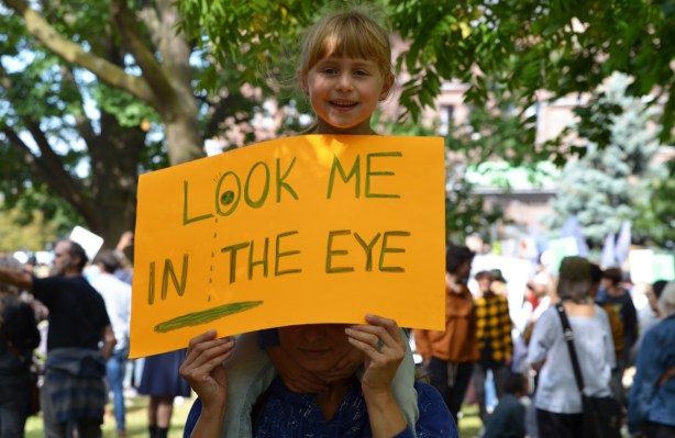 at the climate strike rally at Queens Park on a sunny morning in September, a girl is on her parent's shoulder. she is holding a yellow sign that says look me in the eye 