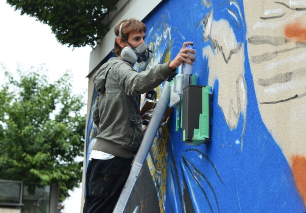 a man, street artist Mateo, spray paints a mural as he stands on a ladder. He's wearing a respirator, painting with blue paint