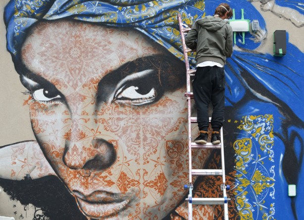 a man is on a ladder as he paints a mural of a woman with henna coloured stencils all over her face, and a blue and yellow head scarf, 