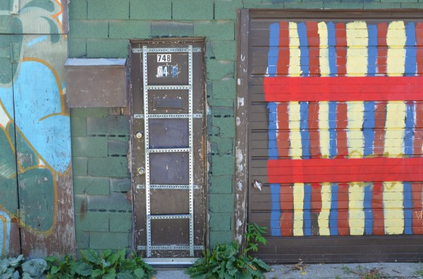 a narrow brown door with metal strapping grid on it beside a garage door with red, yellow, and blue stripes, wall is covered with green shingles 