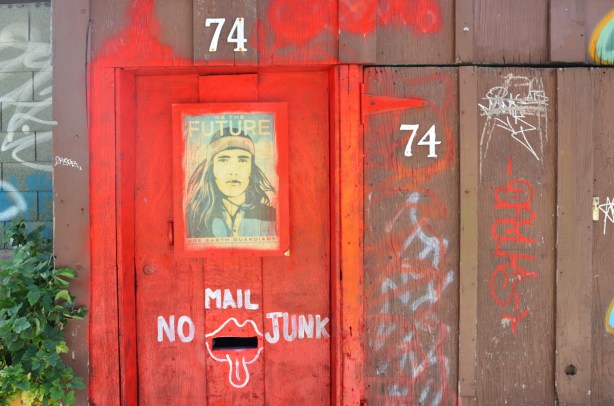 a wood wall and door in an alley painted red and brown, the number 74 on it twice, a mail slot with white paint around it to make slot look like mouth with tongue stuck out, no junk mail written too, a picture of a man on the door with the words we the future