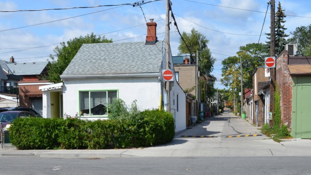 a very small white house at the corner of a street and an alley, a view up the alley 