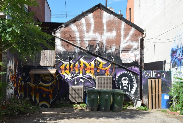 back of a building, in an alley, covered with street art. On top is a large white orek in white, green garbage bins in front 