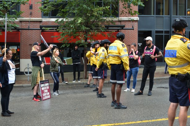 two sides of a protest speaking, while police keep them separated. Christian side is filming the conversation, counter protesters are wearing T shirts that say army of love 