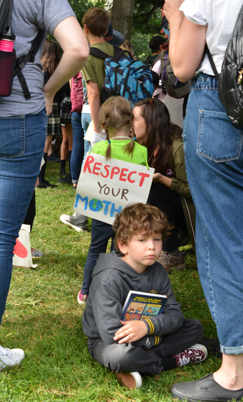 at the climate strike rally at Queens Park on a sunny morning in September, a young boy sits on the grass between two women. Behind him is girl with a sign on her back that says Respect your Mother 