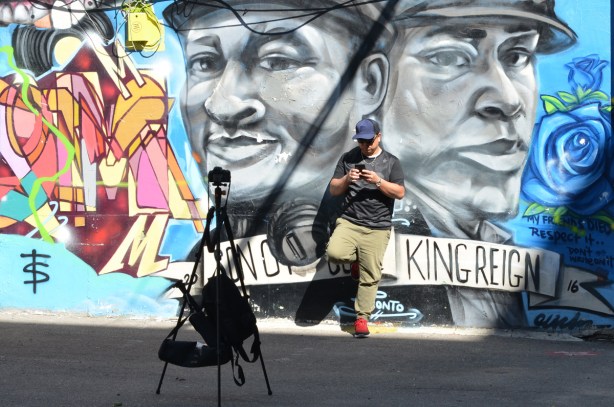 a man has set up a tripod to take a selfie of him positioned between two large faces painted in an alley