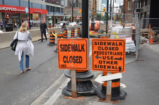 two orange signs saying sidewalk closed, and arrows, barriers on street to form a passage for pedestrians on the street, two people walking by 
