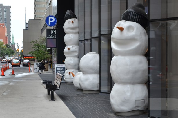 sculptures on sidewalk, snowmen, beside a new condo, one snowman has lost its head 