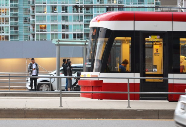 back end of a new TTC streetcar, at a stop on Spadina, people waiting, a man is on his phone as he waits, condo behind 