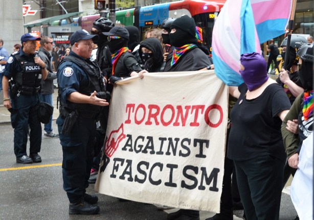 a toronto police officer talks to a group with faces partially covered and holding a banner that says Toronto against fascism 