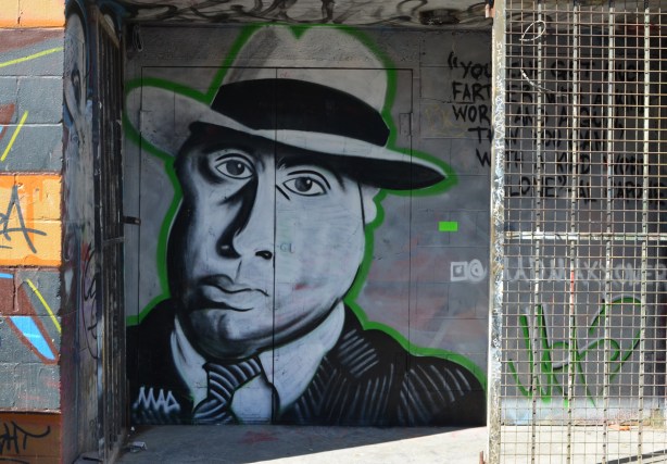 mural in an alley doorway, portrait of Al Capone in shades of grey, wearing a fedora, accompanied by words 