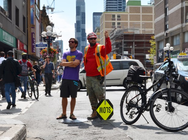 two men standing beside a police bike on the street. on the right, a construction workman with orange reflective vvest, holding a slow sign upside down and on the left, a young man in a purple t shirt and dark shorts, watching a protest on Yonge street