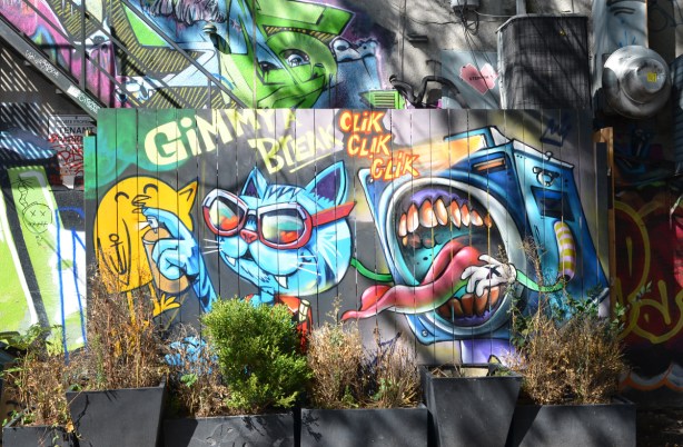 street art by uber5000, a yellow birdie, a blue cat and a characterization of a camera as a face with large mouth and sharp teeth, also with long tongue sticking out. words say gimme a break clik clik clik