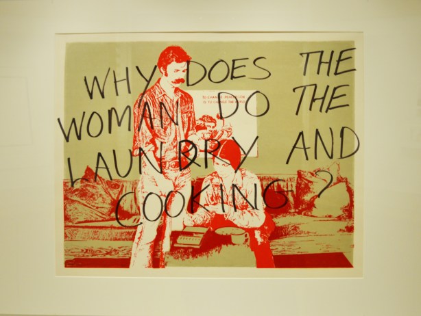 q print that shows a woman working, in red ink on green background, with black words written on top of it, why does the woman do the laundry and cooking
